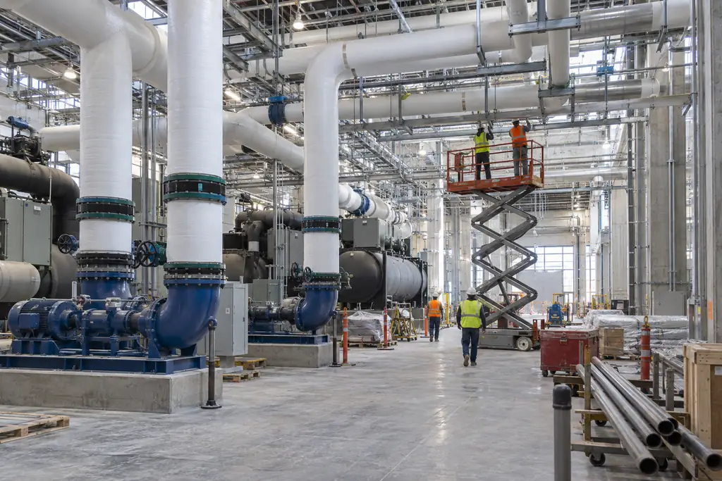 Crew on a scissor lift installing pipe supports above chillers, pumps and insulated chilled water pipe in a central utility plant under construction