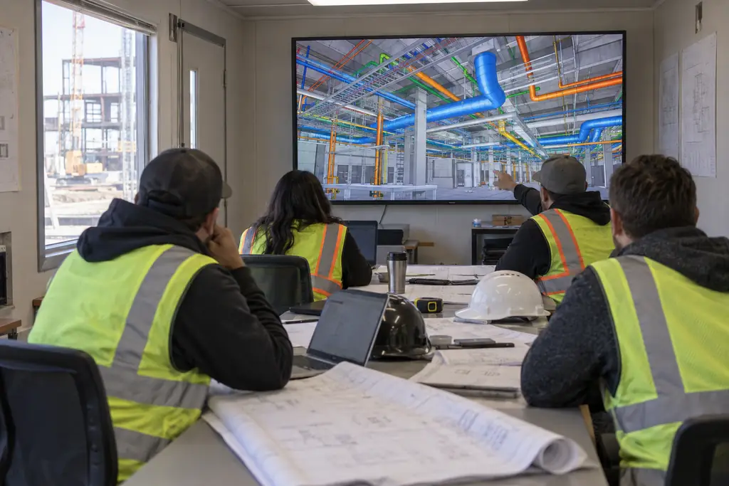 Four people in high-visibility vests in a job trailer looking at a 3D model of ducts and pipes on a large screen