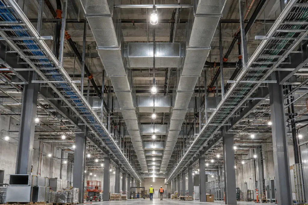 Overhead ductwork, cable trays and pipe racks in an unfinished data hall ceiling, with two workers walking below